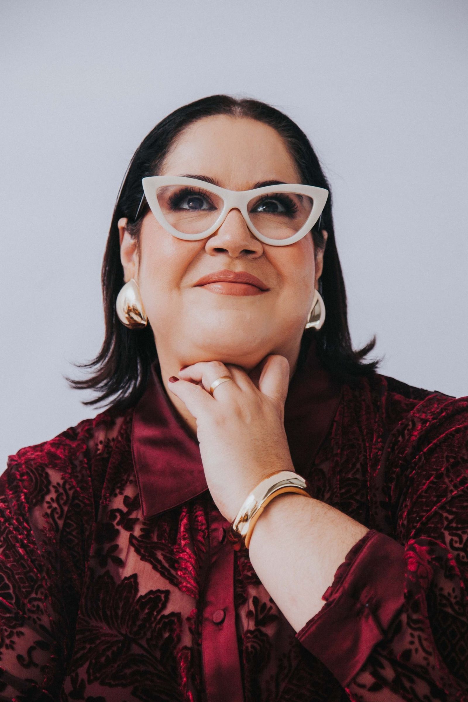 Retrato de Viviane Veiga Távora, de óculos claros e camisa vinho, com a mão no queixo e o olhar voltado para o alto